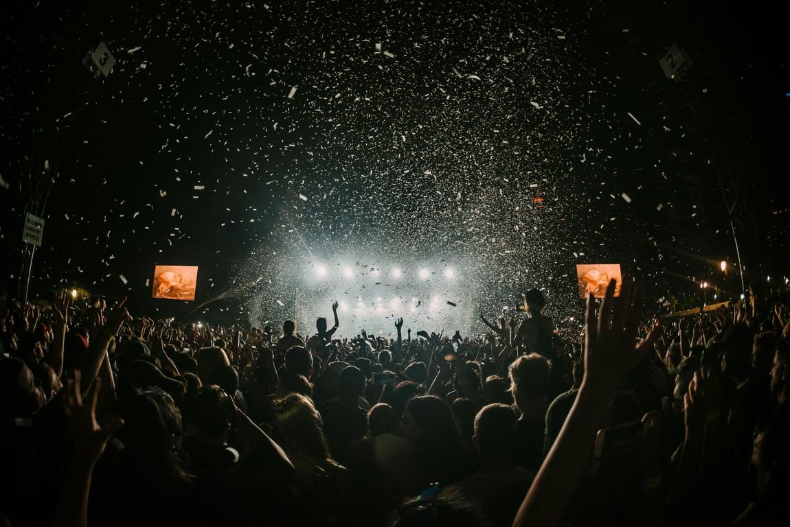 A packed festival crowd at night, hands in the air, confetti falling in front of a brightly lit main stage