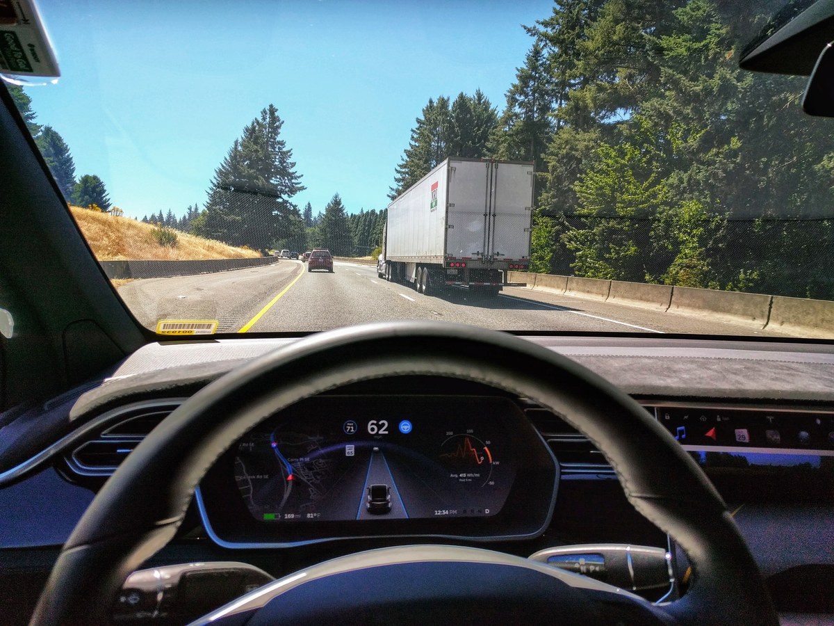The view from the driver's seat of a Tesla Model X on a highway, hands off the wheel, with the instrument cluster showing the car tracking its lane behind a semi truck