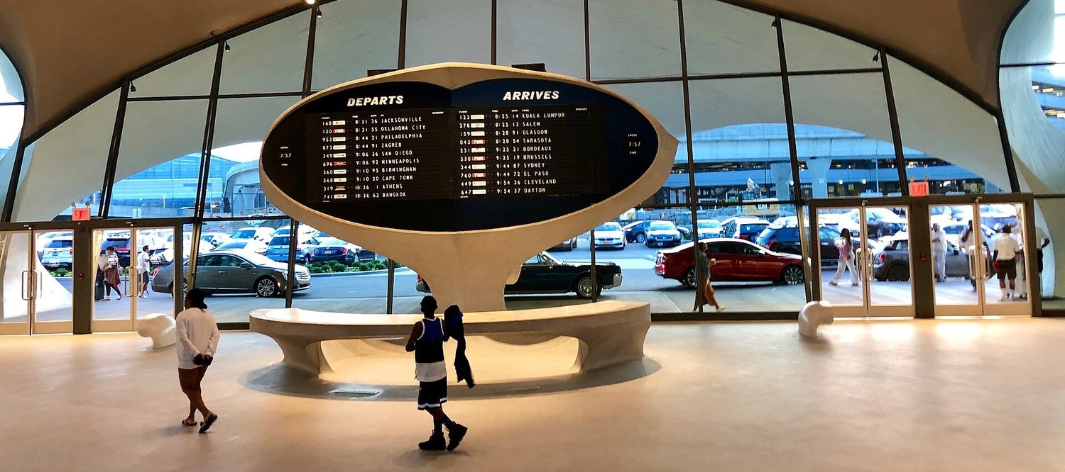 The split-flap departures and arrivals board in the TWA Flight Center at JFK airport, with travelers walking past.