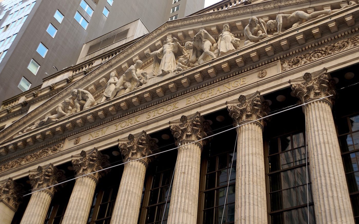 Upward view of the New York Stock Exchange's stone facade on Broad Street, with tall fluted columns, sculpted figures in the pediment and &ldquo;NEW YORK STOCK EXCHANGE&rdquo; lettering.